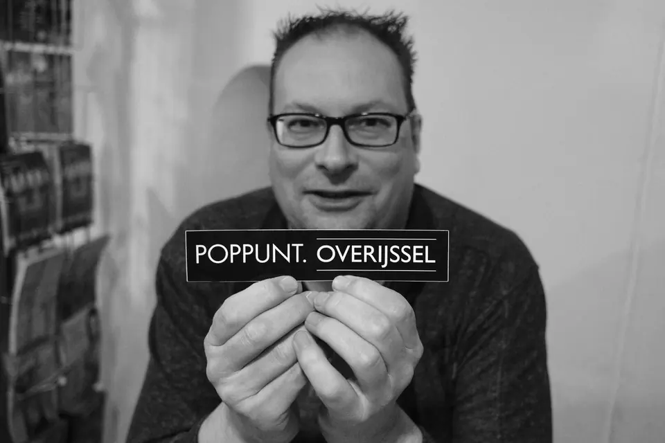 Zwart-witportret van een man met bril die een bordje met Poppunt Overijssel vasthoudt
