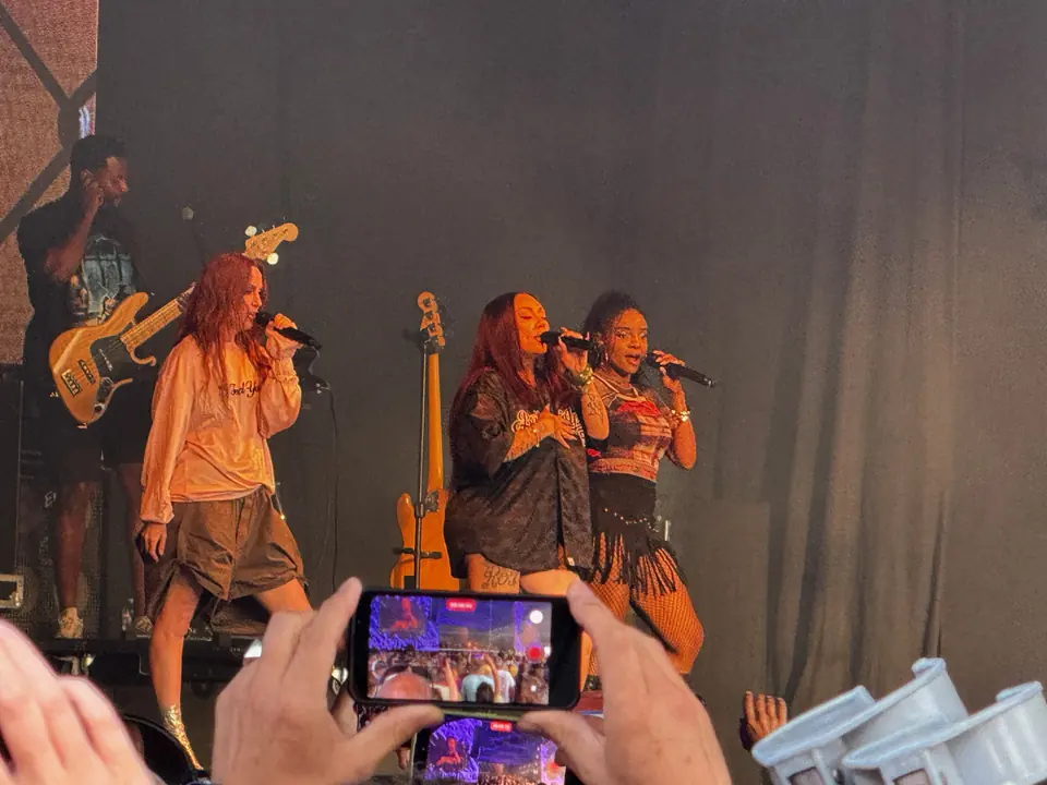 Drie zangeressen met een bassist op het podium van Dauwpop, met een telefoon op de voorgrond