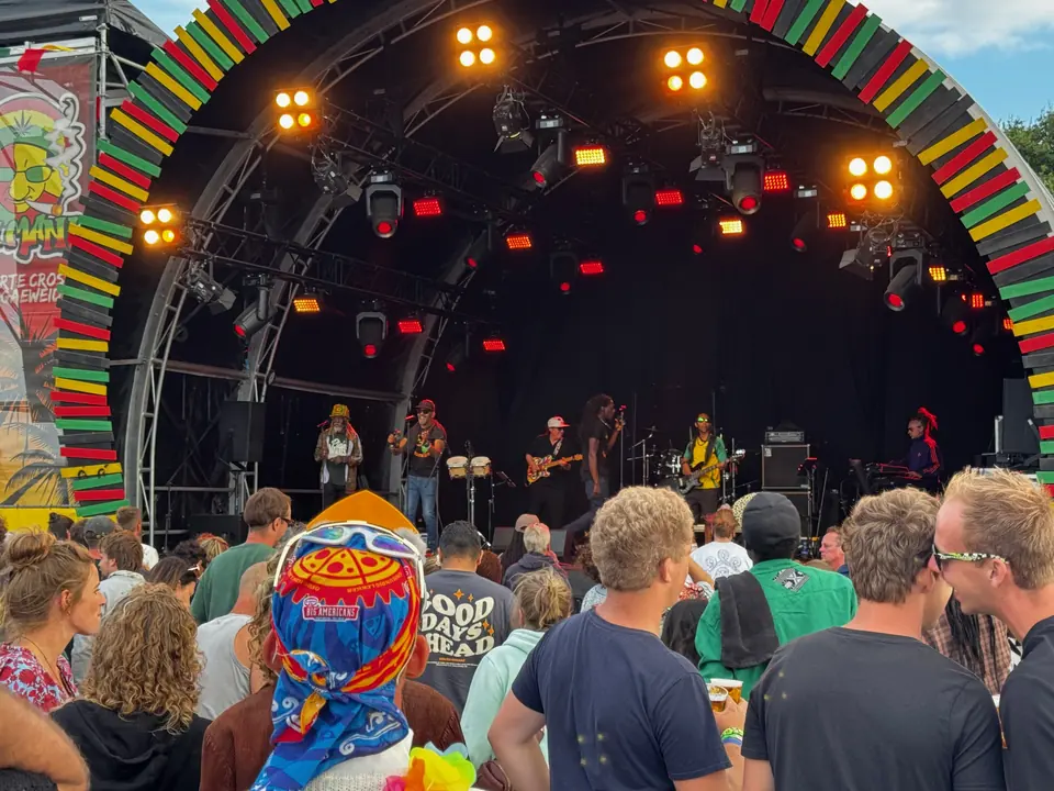 Een podium met een regenboogboog in reggaekleuren en publiek met kleurrijke hoeden ervoor